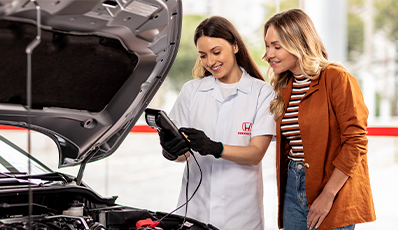 Consultora técnica da Honda mostra o resultado de um diagnóstico veicular para uma cliente, ambas sorrindo ao lado de um carro com o capô aberto dentro da concessionária.